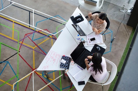 Two people sitting at a table with laptops, talking